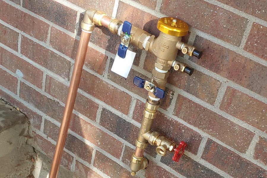 New backflow preventer plumbed in copper against a brick wall above a fresh trench